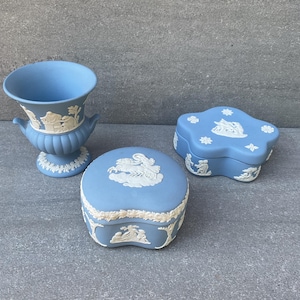 Puede incluir: Tres piezas decorativas de cerámica azul claro. Un jarrón con asas y base redonda, y dos cajas con tapa. Todas las piezas presentan diseños en relieve blancos de figuras clásicas y motivos florales. Las piezas están dispuestas sobre una superficie gris.