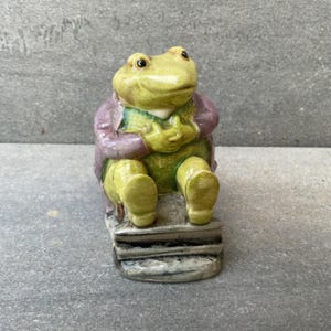 May include: A ceramic frog figurine, seated and wearing a purple jacket over a green vest and shirt. The frog has yellow-green skin and is sitting on a stack of books. The figurine has a whimsical, storybook aesthetic.