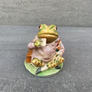 Könnte beinhalten: Eine Keramikfigur eines Frosches, der ein rosa-violettes Outfit trägt und auf einem grünen Sockel sitzt. Der Frosch hat einen grün-gelben Körper mit braunen Flecken und hält ein kleines weißes Objekt in seiner Hand.