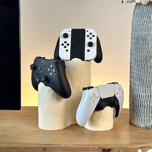 May include: Three different video game controllers are displayed on a white, textured stand. The controllers are black, white, and gray. The stand is on a wooden surface. The background includes a vase with white cotton flowers and a black television.