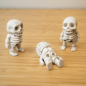 May include: Three white plastic skeleton figurines. Two are standing upright, while the third is lying on its back.