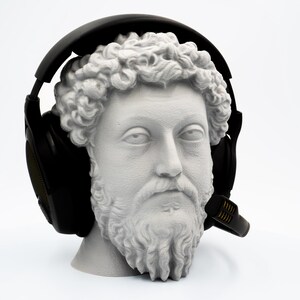 May include: A white bust of a man with a beard and curly hair wearing black headphones with a microphone. The bust is set against a white background.