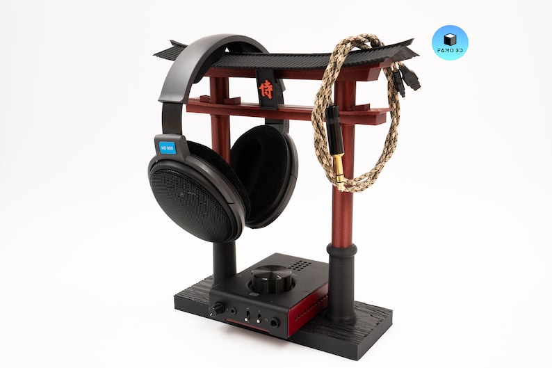 May include: A headphone stand in the shape of a Japanese torii gate, with a black base and red accents. A pair of black headphones and a braided cable are displayed on the stand. The stand also holds a black and red audio amplifier.