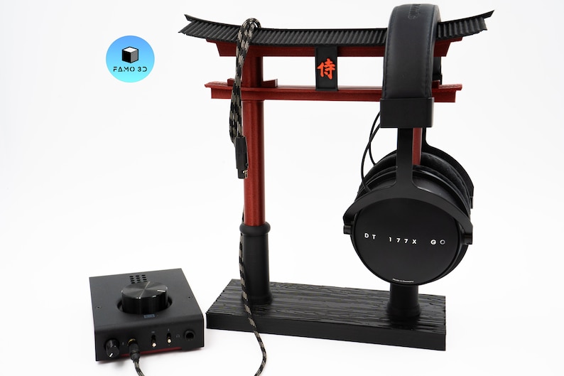 May include: A black and red headphone stand shaped like a Japanese torii gate, holding black headphones. A black audio amplifier sits to the left. The headphones are labeled "DT 177X GO". The stand has the text "侍" on it.