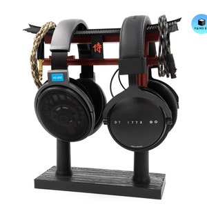 May include: A black headphone stand with two pairs of headphones. The stand has a red and black torii gate design. One pair of headphones is black with "HD 600" on the side, and the other is black with "DT 177X GO" on the side.