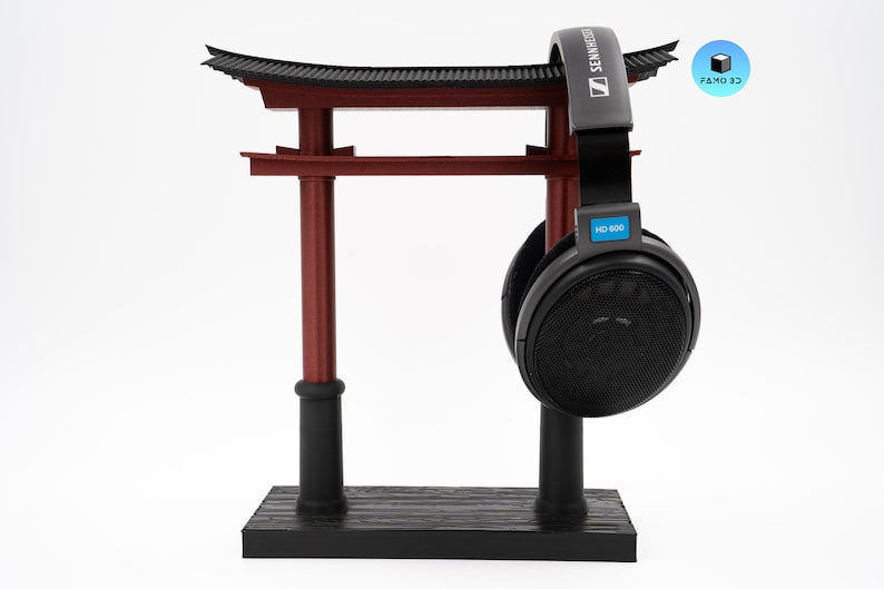 May include: A 3D-printed headphone stand shaped like a Japanese torii gate, in red and black. A pair of black Sennheiser HD 600 headphones hangs from the top. The stand is on a white background.