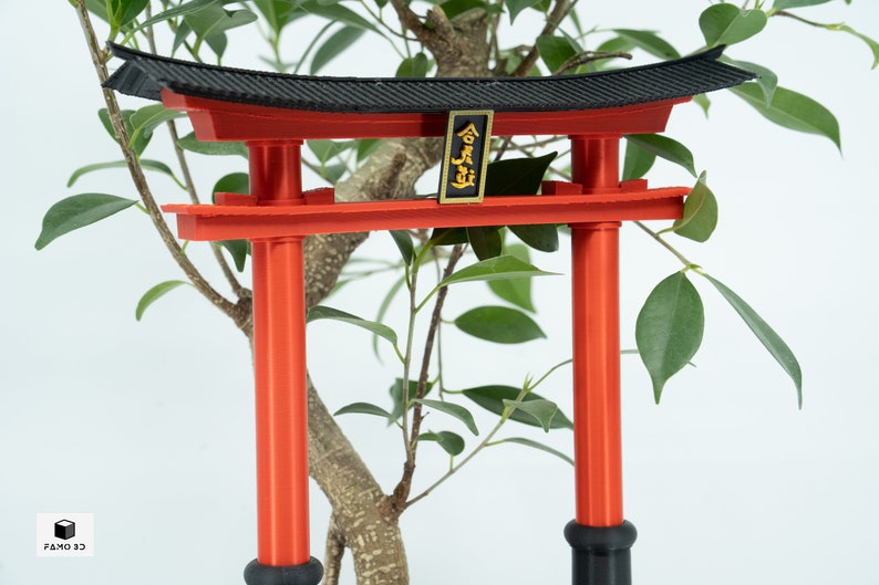 May include: A red and black miniature torii gate with a gold plaque that reads "合掌" in Japanese characters. The gate is surrounded by green leaves.