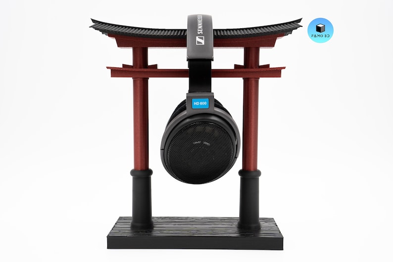 May include: A black and red headphone stand shaped like a Japanese torii gate, holding a pair of black headphones. The headphones have the text "HD 600" on them. The stand has the logo "FAMO 3D" on it.