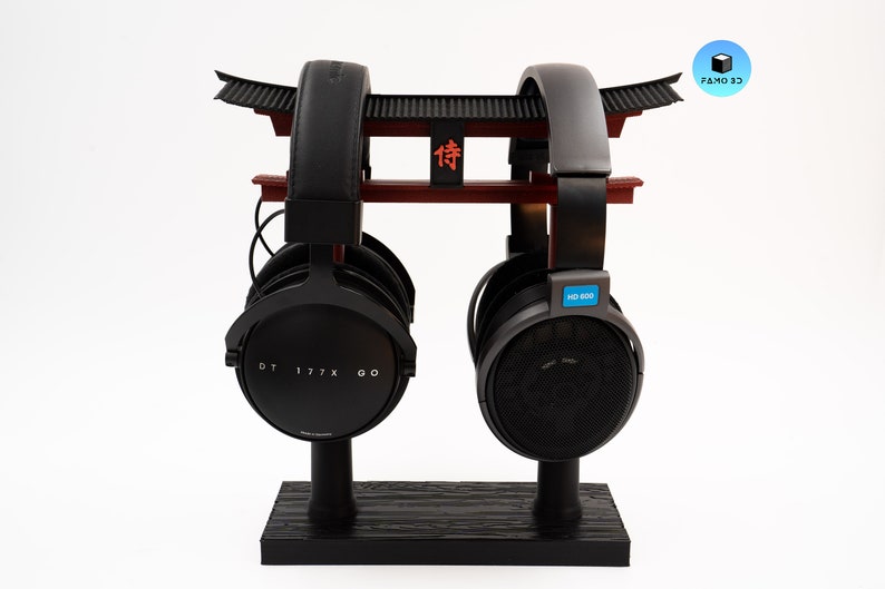 May include: A black headphone stand shaped like a Japanese torii gate, holding two pairs of headphones. The stand is black with red accents and the word "侍" in red. One pair of headphones is labeled "DT 177X GO" and the other "HD 800".