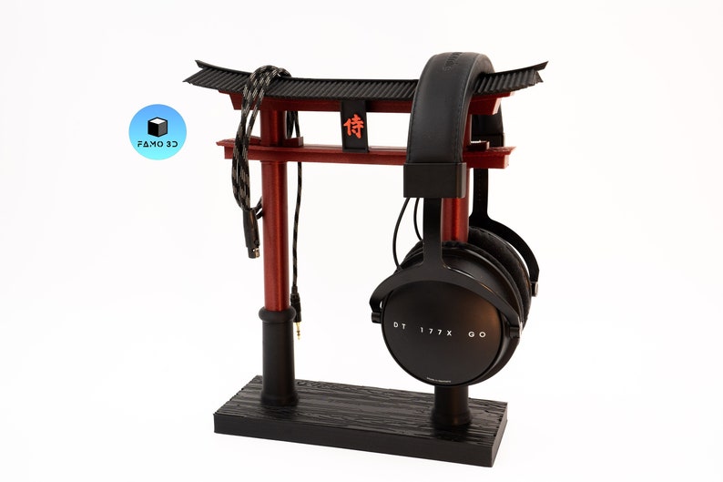 May include: A black and red headphone stand shaped like a Japanese torii gate. The stand holds a pair of black headphones with the text "DT 177X GO". A black and gray cable is draped over the top of the gate.