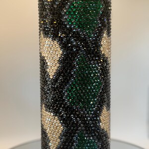 May include: A bedazzled water bottle with a black lid. The bottle is covered in black, green, and gold rhinestones in a camouflage pattern. The bottle is cylindrical and appears to be made of stainless steel.