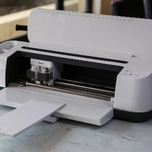 May include: A white Cricut Explore Air 2 cutting machine with the lid open and a white mat extended. The machine has a black base and a silver cutting blade.
