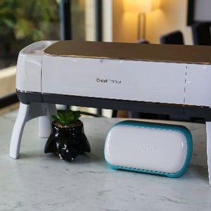 Puede incluir: Una máquina de corte Cricut Maker blanca con una parte superior plateada y una base negra. La máquina está sobre un soporte blanco con cuatro patas. Una pequeña Cricut EasyPress blanca y turquesa está delante de la máquina.