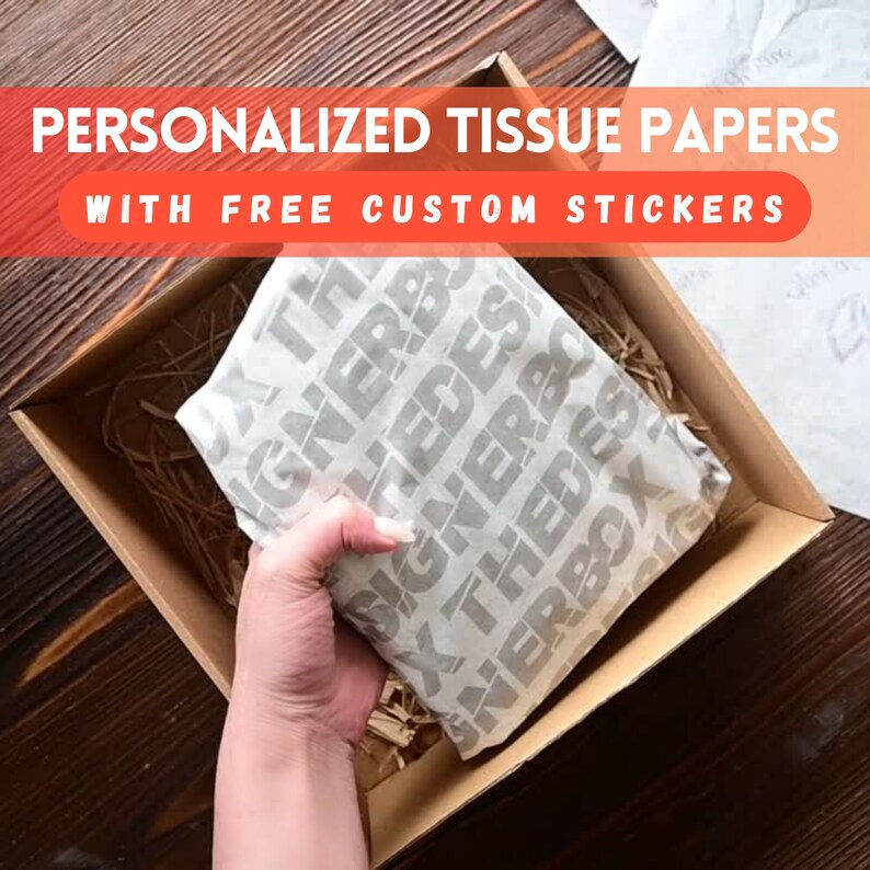 May include: A brown cardboard box with a white tissue paper inside. The tissue paper has a repeating pattern of the words "The Box" in a gray font. The box is sitting on a wooden surface. The text on the box reads "Personalized Tissue Papers with Free Custom Stickers".