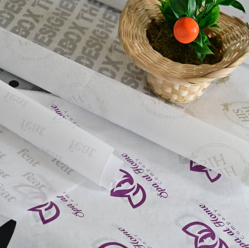 May include: White tissue paper with a purple logo that says "Spa at Home Company" and the text "His" in a circle. The tissue paper is rolled up and there is a small potted plant with an orange fruit in the background.