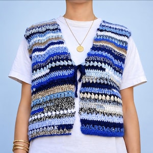 Op de afbeelding: Een mouwloos gehaakt vest met horizontale strepen in diverse tinten blauw, wit en beige. Het kledingstuk heeft een sluiting aan de voorzijde met strikbandjes van bijpassend blauw garen en wordt gedragen over een effen wit t-shirt.