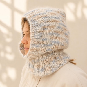 May include: A knitted balaclava hood featuring a variegated yarn pattern in soft shades of beige, cream, and light blue. The headpiece includes a ribbed neck section and a structured hood opening with a matching ribbed trim along the edge.