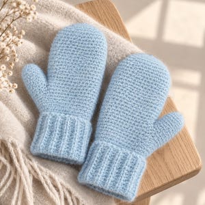 May include: A pair of light blue mittens crafted with a textured crochet stitch. Each mitten features a distinct thumb section and a ribbed cuff at the wrist. The yarn has a soft, slightly fuzzy appearance, providing a consistent solid color throughout.