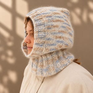 May include: A knitted balaclava hood featuring a variegated yarn pattern in soft shades of light blue, beige, and cream. The headpiece has a ribbed texture around the face opening and a wide ribbed neck section that sits comfortably around the collar.