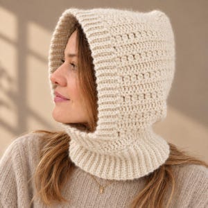May include: A cream-colored knit balaclava hood featuring a ribbed trim along the face opening and a textured stitch pattern throughout the main body. The headpiece extends down to cover the neck with a ribbed collar base in a matching yarn.