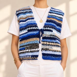 May include: A sleeveless crochet waistcoat featuring horizontal stripes in shades of navy blue, light blue, white, and tan. The waistcoat has a V-neckline with a front tie closure made of matching yarn. It is worn over a plain white t-shirt and white trousers.