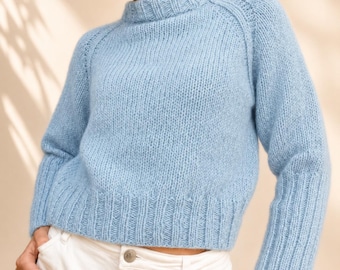 May include: A light blue long-sleeved jumper featuring a chunky knit texture, a ribbed crew neckline, and wide ribbed cuffs at the wrists and waist. The garment is worn with white trousers, showcasing a simple, solid-coloured design with raglan sleeves.
