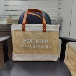 Pu&ograve; includere: Una borsa tote beige con manici in pelle marrone e la scritta "HOLLISTER SANTA BARBARA" in bianco. La parte superiore e inferiore della borsa sono rifinite con tessuto bianco. La borsa &egrave; su una sedia nera.