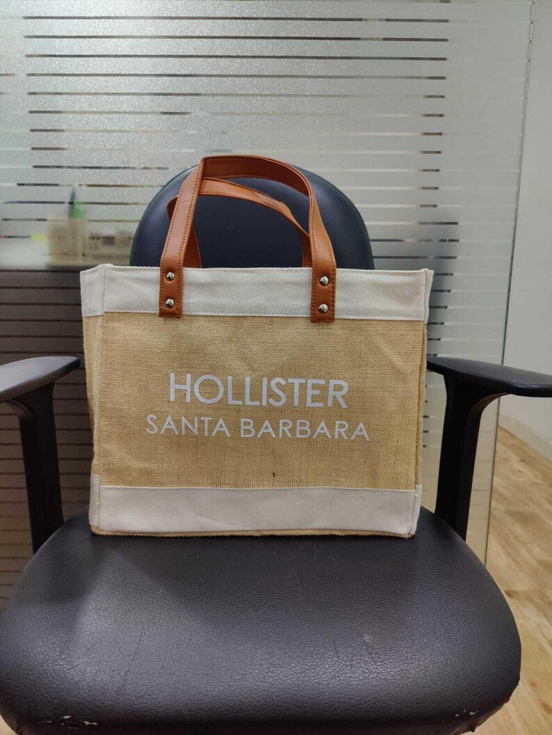 Pu&ograve; includere: Una borsa tote beige con manici in pelle marrone e la scritta "HOLLISTER SANTA BARBARA" in bianco. La parte superiore e inferiore della borsa sono rifinite con tessuto bianco. La borsa &egrave; su una sedia nera.