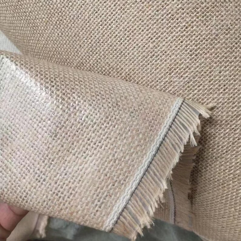 Pu&ograve; includere: Primo piano di un rotolo di tessuto di iuta naturale. Il tessuto ha una trama grossolana e intrecciata di colore marrone chiaro. Il bordo del tessuto &egrave; sfilacciato, rivelando le singole fibre. Il tessuto &egrave; arrotolato, mostrando l'interno e l'esterno.