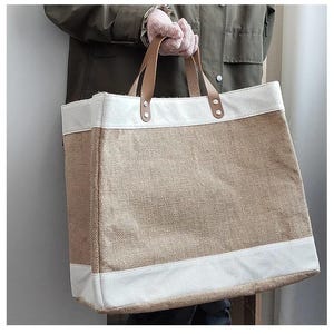 Pu&ograve; includere: Una grande borsa tote con un corpo in juta naturale e finiture in tela bianca. La borsa presenta manici in pelle marrone fissati con rivetti metallici. La borsa &egrave; tenuta da una persona che indossa una giacca verde e guanti rosa.