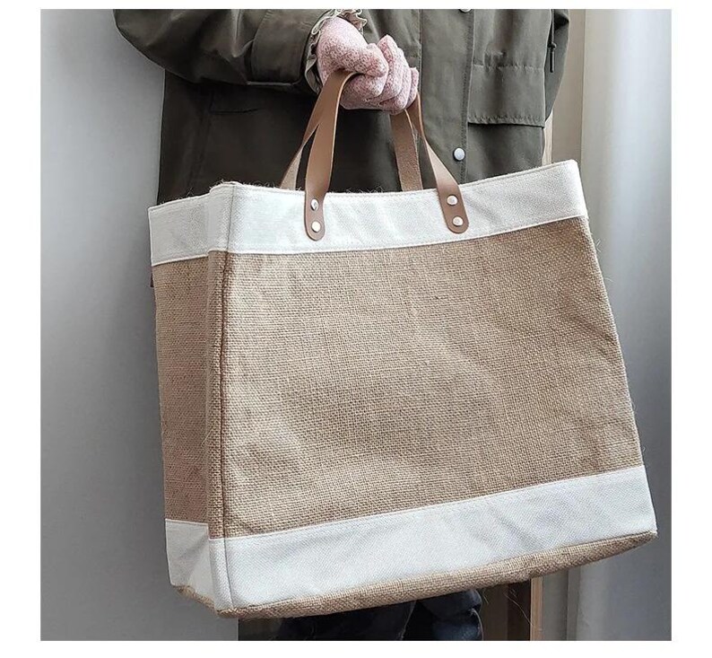 Pu&ograve; includere: Una grande borsa tote con un corpo in juta naturale e finiture in tela bianca. La borsa presenta manici in pelle marrone fissati con rivetti metallici. La borsa &egrave; tenuta da una persona che indossa una giacca verde e guanti rosa.
