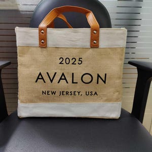 Pu&ograve; includere: Una borsa tote beige con la scritta "2025 AVALON NEW JERSEY, USA" in nero. La borsa ha un bordo bianco superiore e inferiore e manici in pelle marrone. La borsa &egrave; appoggiata su una sedia nera.