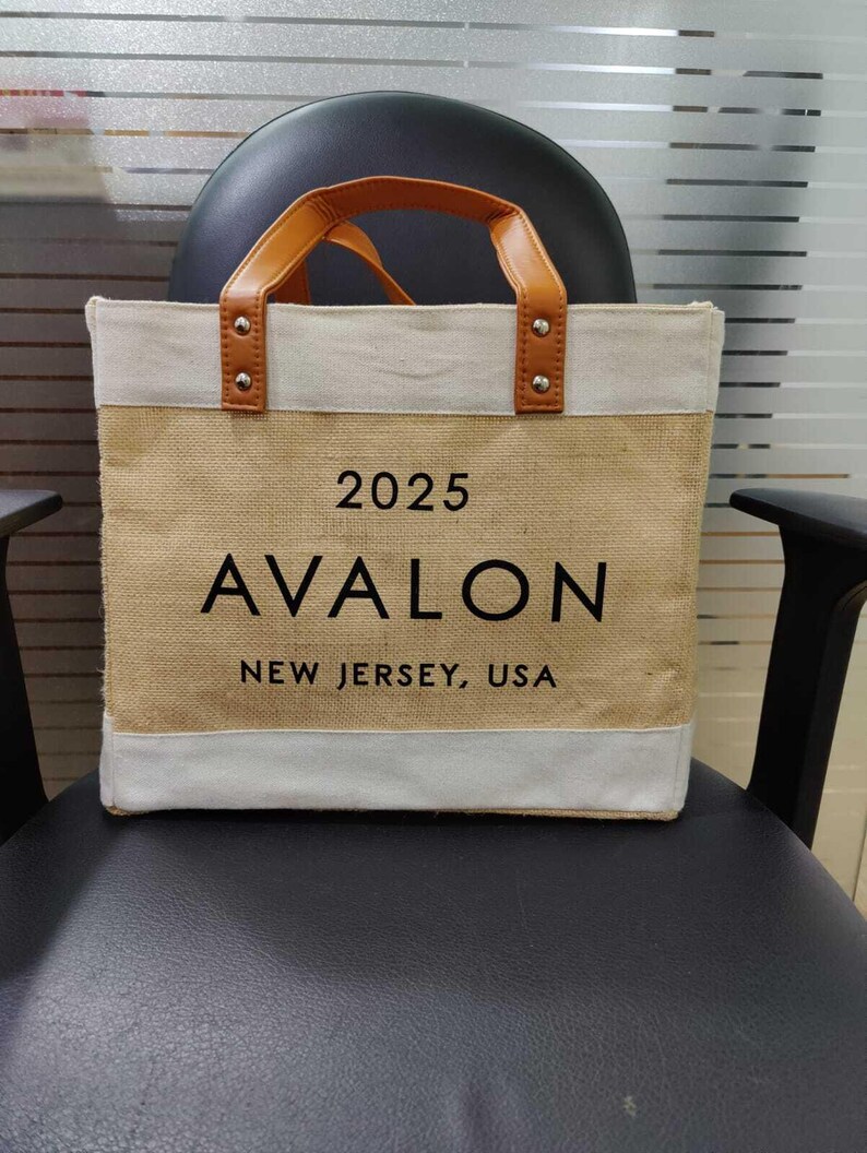 Pu&ograve; includere: Una borsa tote beige con la scritta "2025 AVALON NEW JERSEY, USA" in nero. La borsa ha un bordo bianco superiore e inferiore e manici in pelle marrone. La borsa &egrave; appoggiata su una sedia nera.