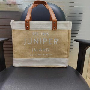 Pu&ograve; includere: Una borsa tote beige con manici in pelle marrone e finiture bianche. La borsa presenta la scritta "EST. 1895 JUNIPER ISLAND STONY LAKE, ONTARIO" in lettere bianche. La borsa &egrave; appoggiata su una sedia nera.