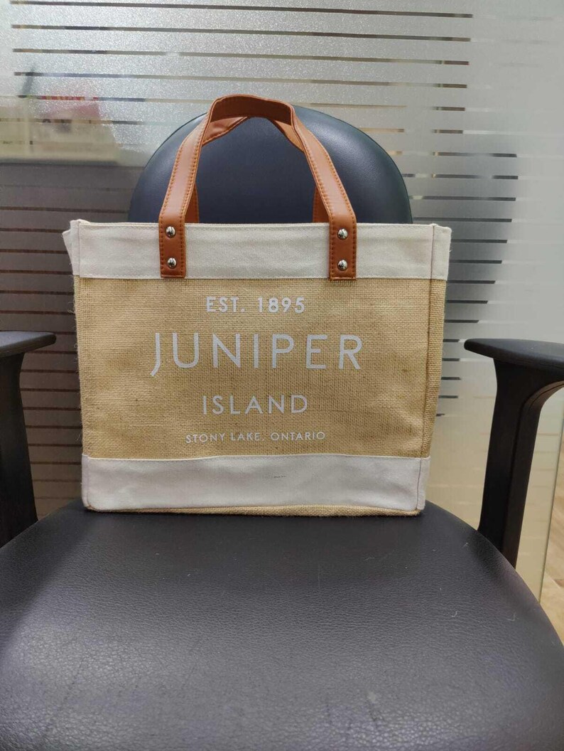 Pu&ograve; includere: Una borsa tote beige con manici in pelle marrone e finiture bianche. La borsa presenta la scritta "EST. 1895 JUNIPER ISLAND STONY LAKE, ONTARIO" in lettere bianche. La borsa &egrave; appoggiata su una sedia nera.
