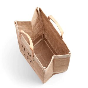 Pu&ograve; includere: Una borsa tote rettangolare in iuta con manici in pelle marrone chiaro. La borsa ha le parole "you are" stampate in nero sul davanti. La borsa &egrave; vuota e si trova su una superficie bianca.