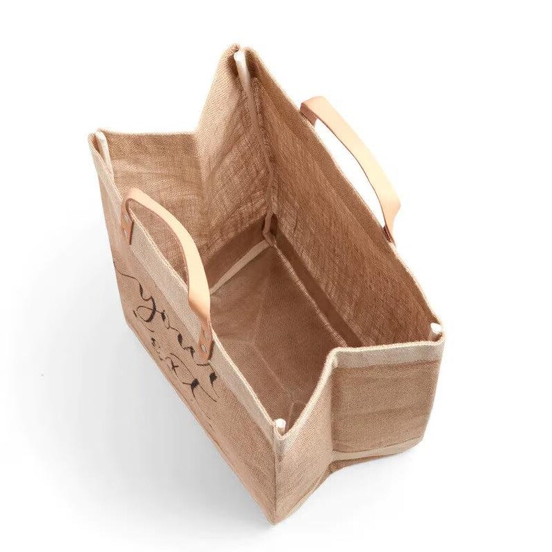 Pu&ograve; includere: Una borsa tote rettangolare in iuta con manici in pelle marrone chiaro. La borsa ha le parole "you are" stampate in nero sul davanti. La borsa &egrave; vuota e si trova su una superficie bianca.