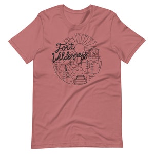 May include: A pink t-shirt with a black graphic design that says "Life is better at Fort Wilderness" with a sun, trees, a tent, a campfire, a lake, a dock, and a water tower.