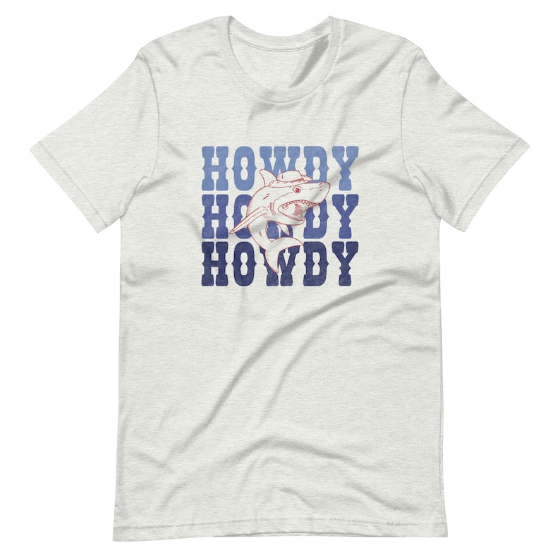 Puede incluir: Una camiseta blanca con un gr&aacute;fico azul y rojo de un tibur&oacute;n con un sombrero de vaquero. El texto "HOWDY" se repite tres veces en un color azul descolorido.