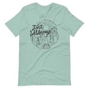 May include: A light blue t-shirt with a black graphic design of a sun, trees, a lake, a dock, a campfire, and a tent. The text "Life is better at Fort Wilderness" is written in a cursive font.