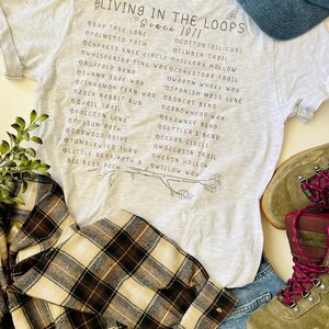 May include: A gray t-shirt with the text "Living in the Loops Since 1971" and a list of trail names printed on it. The shirt is paired with a blue baseball cap and a pair of brown and beige plaid flannel shirt.