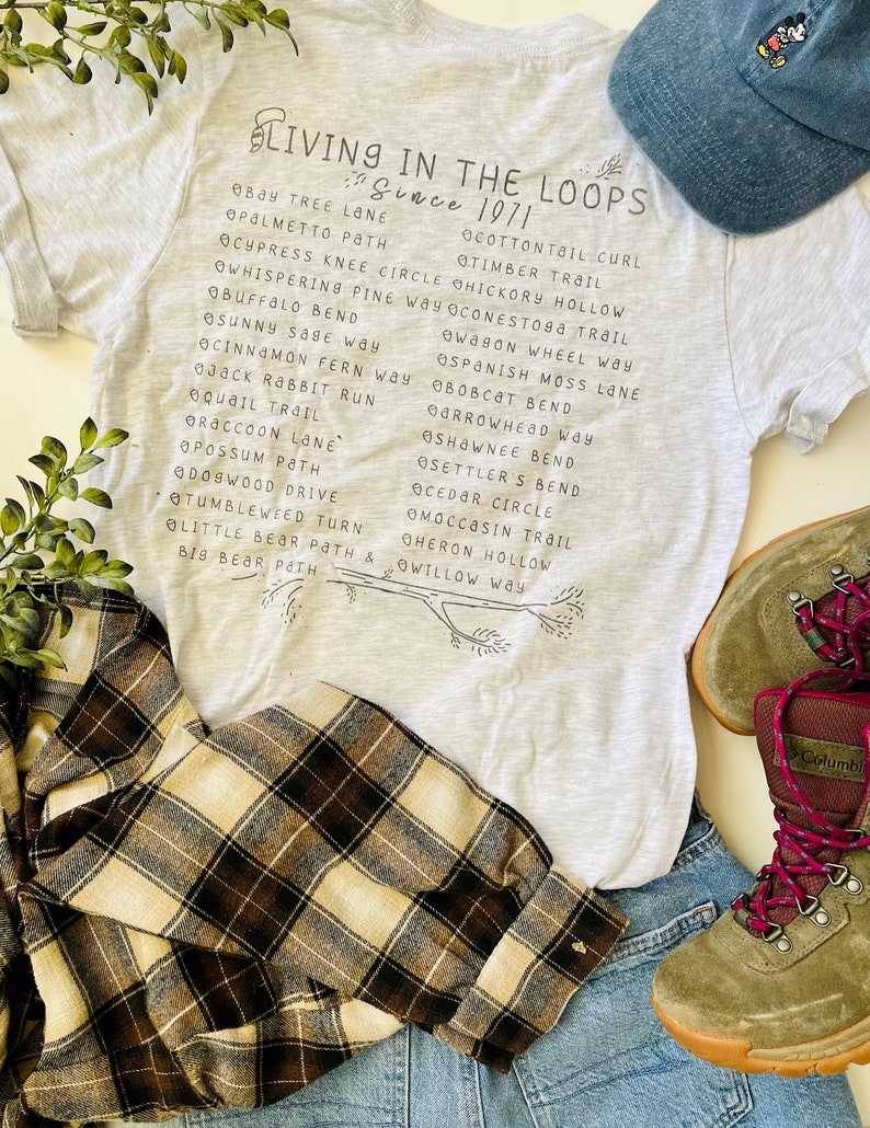May include: A gray t-shirt with the text "Living in the Loops Since 1971" and a list of trail names printed on it. The shirt is paired with a blue baseball cap and a pair of brown and beige plaid flannel shirt.