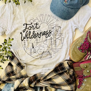 May include: A gray t-shirt with a black graphic design that says "Life is better at Fort Wilderness" with a sun, trees, a lake, a campfire, and a dock. The shirt is paired with a plaid flannel shirt, blue jeans, a blue baseball cap with a Mickey Mouse design, and brown hiking boots with pink laces.
