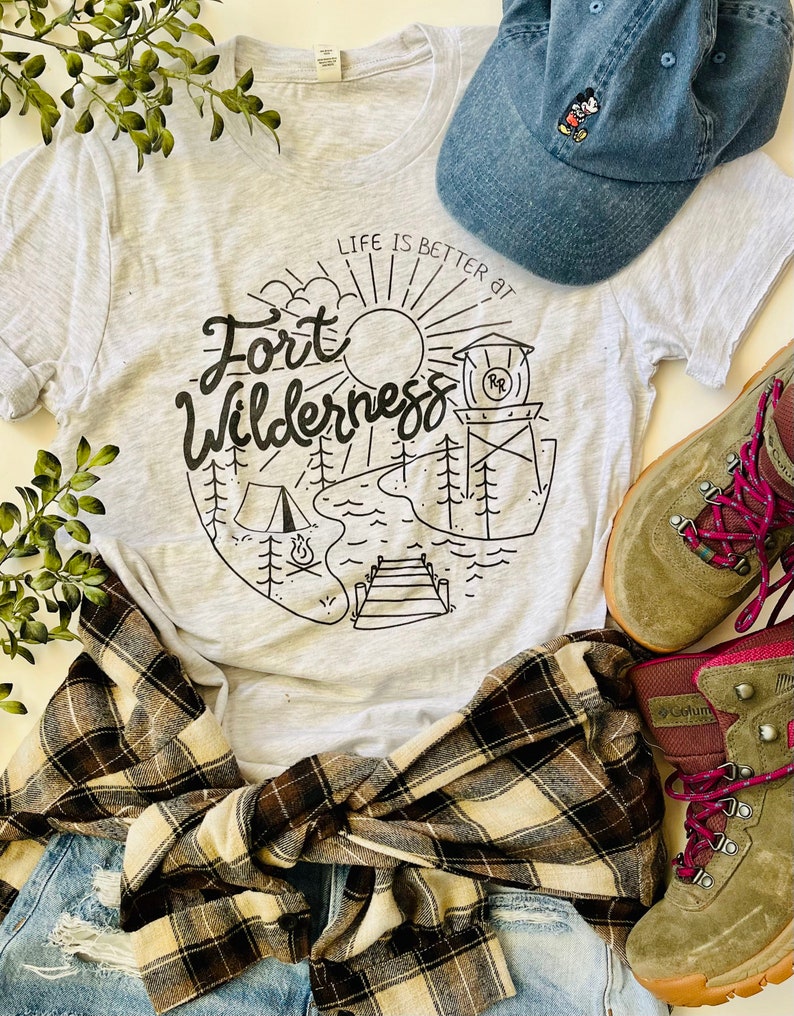 May include: A gray t-shirt with a black graphic design that says "Life is better at Fort Wilderness" with a sun, trees, a lake, a campfire, and a dock. The shirt is paired with a plaid flannel shirt, blue jeans, a blue baseball cap with a Mickey Mouse design, and brown hiking boots with pink laces.