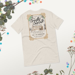 Op de afbeelding: Een beige T-shirt met een grafische print van een winkel genaamd "The Crafty Carver" met een zwarte raaf, een bezem, een mand met paddenstoelen en een beer. De tekst luidt "A Right Mr. Milly The Crafty Carver Maker of High-End, Timed, Carvings and Novelties... Maker of Many a Brighton Bay Rock Chandler, Commemorative Statues & Rare Pages HALF-OFF EVERYTHING  *Goods cannot be divided with partial payments*"