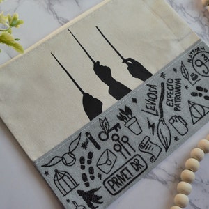 May include: A white canvas zippered pouch with a gray and black print of Harry Potter themed symbols and text, including "Platform 9 3/4", "Expecto Patronum", "LevioSA", and "Privet Dr".