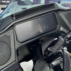 May include: Close-up of a motorcycle dashboard with a large rectangular display screen. The dashboard is black with a clear windshield. The handlebars and controls are visible, with the Harley-Davidson logo on the brake lever.