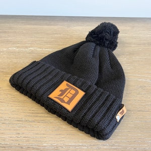 May include: Black knit beanie with a brown leather patch featuring a stylized "D" and a black pom pom on top.