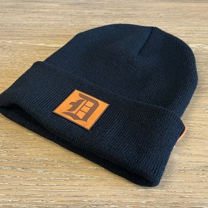 May include: Navy blue knit beanie with a brown leather patch featuring a stylized letter "D". The beanie has a folded brim and a classic, casual style. A great accessory for cold weather.