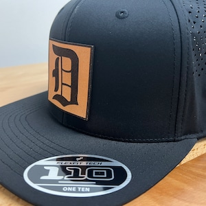 May include: Black baseball cap with a brown leather patch featuring a large black letter "D". The cap has a white logo on the side and a black and white label on the underside of the brim that reads "Flexfit Tech 110 One Ten".